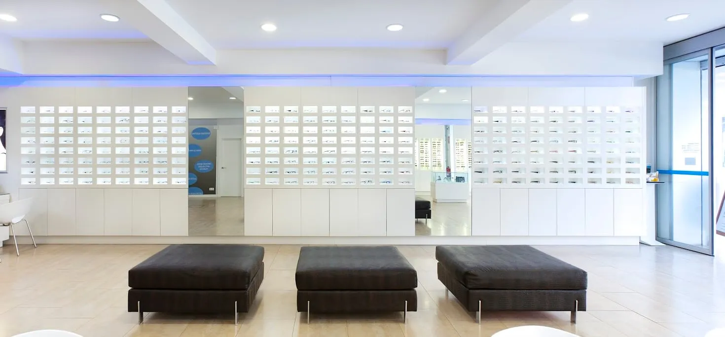 Sala de la óptica con las paredes de monturas iluminadas y tres bancos de espera